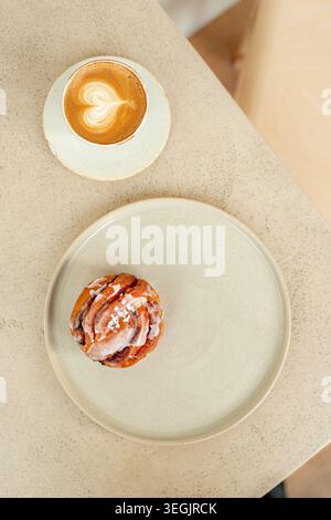 Un colpo sopra la testa di un panino alla cannella fresco smaltato su un piatto di ceramica servito con una tazza di caffè caldo di latte art su un tavolo strutturato. Foto Stock