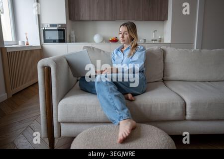 Donna interessata e concentrata, rilassata seduta su un comodo divano con laptop, mentre lavorava in remoto a casa. Concentrati sullo schermo femminile comodamente noi Foto Stock