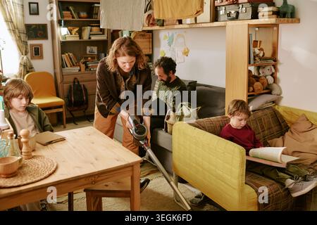 Donna di mezza età che aspira mentre la famiglia si rilassa in una piccola casa Foto Stock