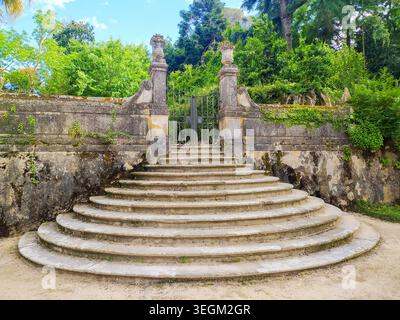 Coimbra, Portogallo - 15 maggio 2026: Storica scalinata semicircolare in pietra che conduce ad un cancello di ferro ornato fiancheggiato da pilastri intemprati in una lussureggiante botanica Foto Stock
