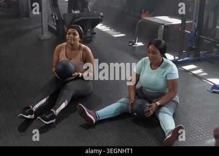 Due donne afroamericane adulte in abbigliamento sportivo che tengono palle mediche e fanno twist sul pavimento della palestra Foto Stock