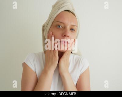 Beauty Woman che tocca delicatamente le guance lisce con entrambe le mani cura della pelle Ritratto con asciugamano turbante, risultato del trattamento facciale domestico Self-Care Wellness. Foto Stock