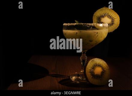 Cocktail kiwi in un bicchiere di coupé con bordo zuccherato e foglie di menta, guarnito con fette di kiwi su un tavolo di legno scuro, cibo da bar in stile umoristico e leggero Foto Stock