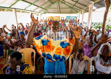 Una donna keniota in colorato abito tradizionale, si erge in una comunità e guida il gruppo in una gioiosa canzone swahili. Foto Stock