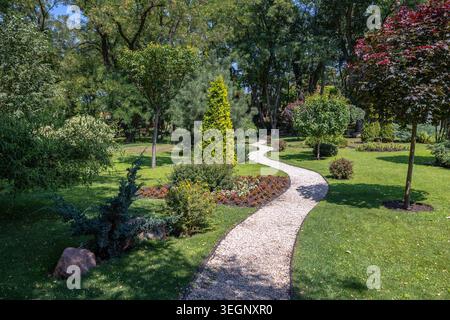 Un tortuoso sentiero di ghiaia si snoda attraverso un giardino ben curato, pieno di prati verdi, alberi diversi, varie conifere ornamentali, e. Foto Stock