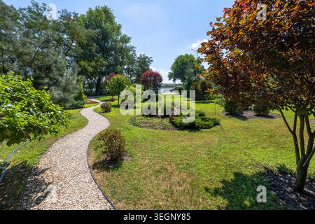 Un tortuoso sentiero di ghiaia si snoda attraverso un parco pubblico paesaggistico con prati verdi, arbusti ornamentali ben curati e vari decidui e c Foto Stock