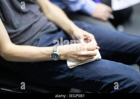 Primo piano di una persona seduta e in mano una penna mentre scrive appunti in un piccolo quaderno a spirale appoggiato sul grembo durante una conferenza o una presentazione Foto Stock