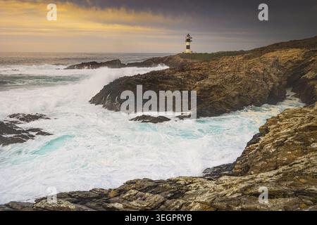Vista del faro di Illa Pancha che si erge su aspre scogliere mentre le potenti onde si infrangono contro le rocce sotto un cielo dorato al tramonto Lugo, Lugo, Spagna. Foto Stock