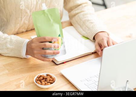 Una persona siede a una scrivania, tenendo in mano un pacchetto verde e bianco di gelatina aromatizzata agli agrumi davanti a un computer portatile aperto e a una ciotola di mandorle. Foto Stock