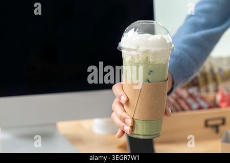 Una persona che tiene in mano un latte matcha alto ghiacciato in una tazza di plastica trasparente con un manicotto di carta, ricoperto di panna montata. Foto Stock