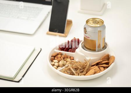 Un piatto di spuntini, tra cui frutta secca, frutta secca e cracker, è disposto su una scrivania accanto a un computer portatile, telefono e una lattina di birra. Foto Stock