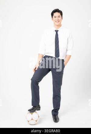 Un giovane asiatico con una camicia bianca, cravatta blu e pantaloni scuri sorride su un pallone da calcio. Foto Stock