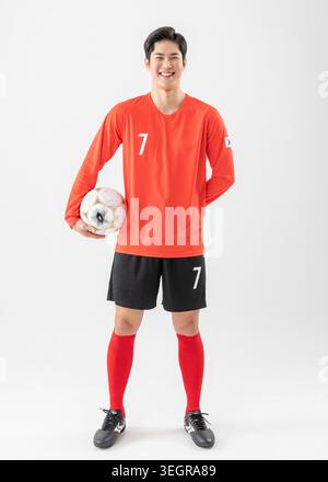 Un giovane giocatore di calcio maschile è in piedi con fiducia, tenendo un pallone da calcio, su uno sfondo bianco. Foto Stock