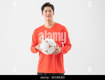 Un giovane calciatore asiatico si erge su un semplice sfondo bianco, reggendo un pallone da calcio e sorridendo. Foto Stock