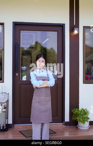 Il personale di una giovane donna asiatica nel grembiule in piedi all'entrata del caffè accoglie i clienti per il servizio di ospitalità Foto Stock