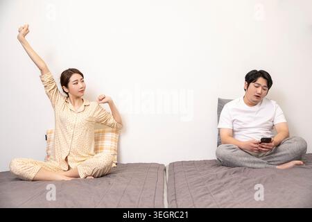 Una donna in pigiama si allunga mentre un uomo si siede sul letto, fissando il suo telefono. Foto Stock