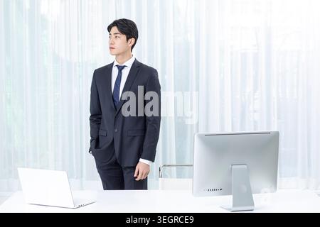 Un giovane uomo d'affari asiatico si trova con fiducia in un ufficio moderno, guardando fuori dalla finestra. Foto Stock