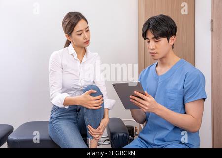 Una giovane donna con una camicia bianca è seduta su un divano nello studio di un medico, le tiene il ginocchio e guarda un tablet con un medico. Foto Stock