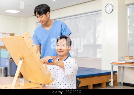 Una giovane infermiera asiatica maschile assiste un paziente asiatico anziano con un esercizio di riabilitazione in una stanza di terapia ospedaliera. Foto Stock