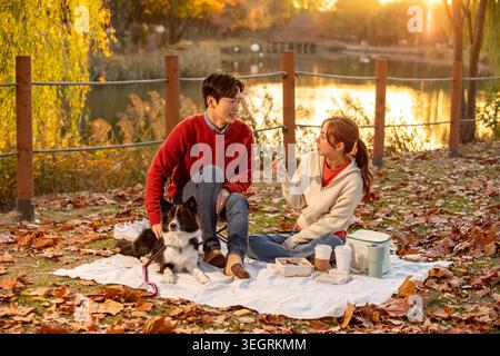 Una giovane coppia si diverte a fare un picnic con il proprio cane in un parco panoramico durante una calda giornata autunnale. Foto Stock