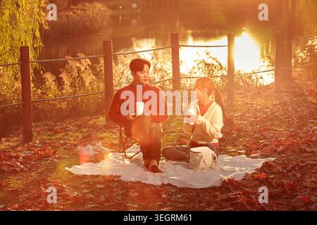 Due giovani adulti si siedono insieme su una coperta da picnic, sorseggiando un caffè e godendosi la calda mattina autunnale. Foto Stock