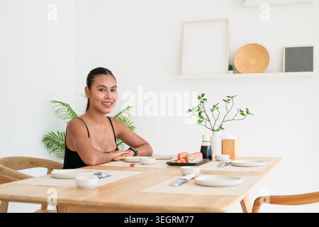 Un'autentica foto orizzontale senza ritocchi di un'adorabile giovane donna tailandese della regione di Isan dei vent'anni, sorridente alla macchina fotografica mentre si siede a una ligh Foto Stock