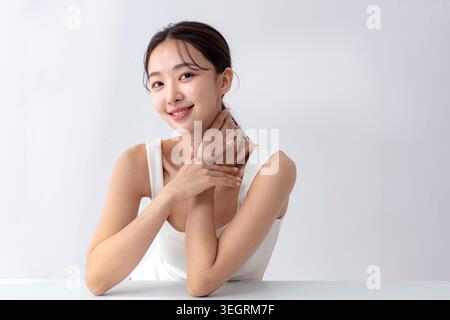 Una giovane donna si siede ad un tavolo bianco, sorridendo e toccando il collo. Foto Stock