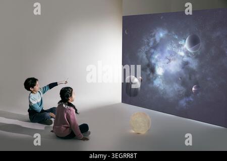 Due bambini seduti sul pavimento, osservando un'immagine proiettata del sistema solare su un grande schermo. Foto Stock