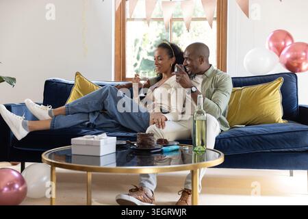 Coppia afroamericana seduta su un divano in velluto blu profondo a casa, tenendo bicchieri da vino e torta Foto Stock