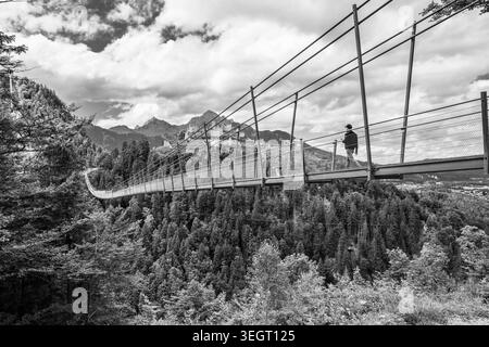 La gente attraversa la Highline 179, un lungo ponte sospeso in Austria. Il ponte si estende su un'ampia gola fluviale e collega le rovine storiche dell'epoca romana. Il paesaggio comprende montagne e alberi. Foto Stock