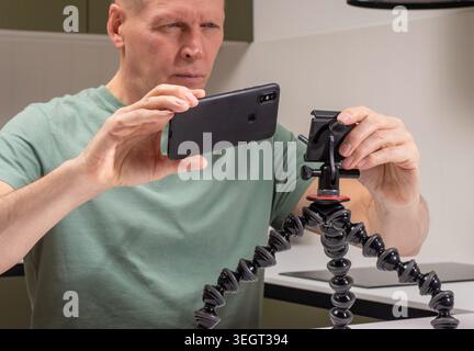 L'uomo maturo posiziona lo smartphone su un supporto per cavalletto flessibile per le riprese. Il maschio adulto installa il dispositivo mobile su un supporto regolabile all'interno del tavolo. Preparati Foto Stock