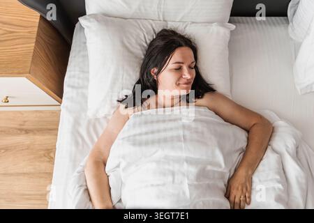 Donna caucasica di mezza età con lunghi capelli scuri giace a letto, sorridendo delicatamente sotto una coperta bianca. La camera ha un design moderno con elementi in legno. Foto Stock