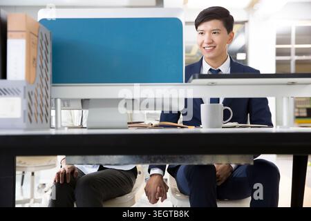 Un giovane asiatico con una tuta da lavoro siede a una scrivania in un ufficio, sorridendo ai suoi colleghi. Foto Stock