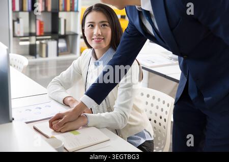 Una giovane donna asiatica con un maglione bianco sorride mentre un uomo adatto con una tuta blu navy la raggiunge per toccare la mano su una scrivania bianca in un ufficio moderno. Foto Stock