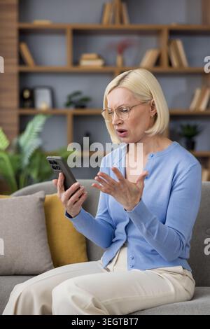 Donna bionda matura che indossa gli occhiali seduti su un divano, gestendo con la mano frustrata mentre guarda uno smartphone, sentendosi stressata e confusa da problemi con la tecnologia Foto Stock