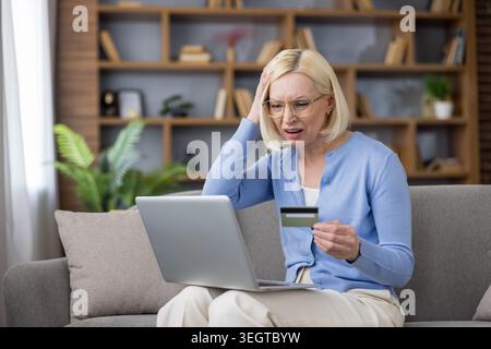 La donna anziana si sente stressata e preoccupata, ha problemi con un pagamento online o ha subito frodi bancarie, ha una carta di credito e utilizza un computer portatile a casa Foto Stock