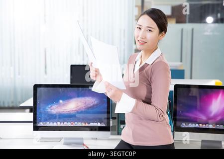 Una donna siede a una scrivania in un ufficio, tenendo una cartella di documenti e sorridendo davanti alla telecamera. Foto Stock