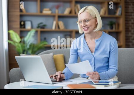 Donna bionda sorridente che indossa gli occhiali seduti su un divano, lavora in remoto dal suo ufficio domestico, utilizza un laptop e tiene in mano documenti cartacei mentre gestisce le attività Foto Stock
