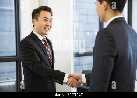 Due uomini d'affari asiatici in tuta che stringono la mano in un ambiente d'ufficio. Foto Stock
