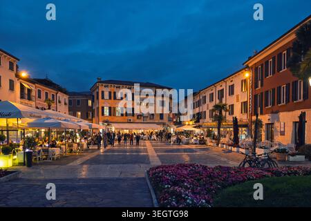Sirmione, Italia - 04.10.2024: Un'incantevole piazza pedonale al crepuscolo, con edifici storici, caffetterie all'aperto e una calda illuminazione stradale. Foto Stock