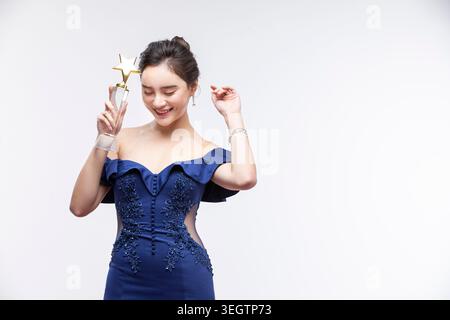 Una giovane donna con un abito formale tiene un trofeo e sorride, ambientato in un contesto neutrale. Foto Stock