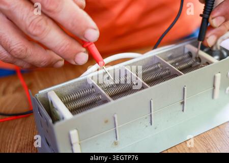 Elettricista adulto specializzato che esegue il test di un componente elettrico utilizzando la mano della sonda del multimetro durante il processo di riparazione controllando il dispositivo di riscaldamento in metallo con una parte profonda Foto Stock