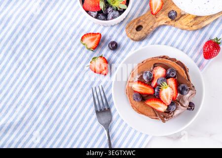 Pila di pancake al cioccolato con una ricca crema di pasta al cioccolato e frutti di bosco freschi - fragole, mirtilli. Colazione dolce e gustosa Foto Stock
