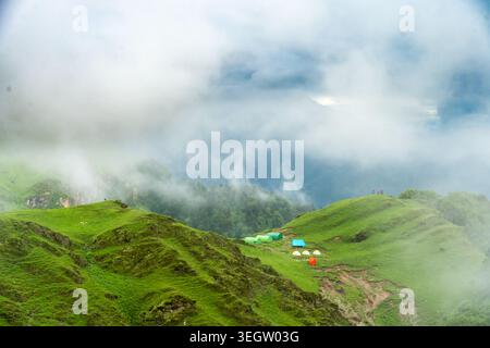 Una vista panoramica di un campeggio di montagna con piccole tende annidate in un vibrante prato verde circondato da una fitta nebbia ad Ali Bedni Bugyal, Chamoli, Uttarak Foto Stock