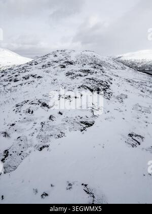 Vista aerea di una cresta di montagna innevata con affioramenti rocciosi sotto un cielo invernale nuvoloso a Ersfjord, Troms, Norvegia. Foto Stock