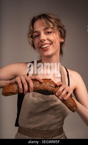 Donna sorridente che indossa un grembiule che rompe una baguette di grano intero, gustando pane caldo fatto in casa Foto Stock
