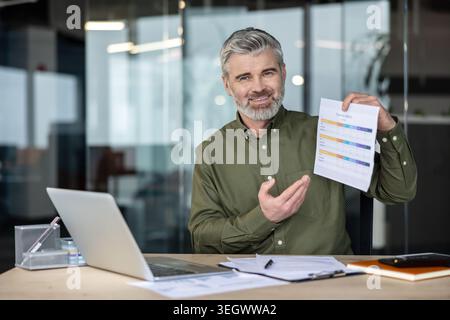 Uomo d'affari maturo e barbuto alla scrivania dell'ufficio, che tiene e indica un rapporto finanziario accanto al suo laptop, sorridendo con fiducia mentre presenta un'analia Foto Stock