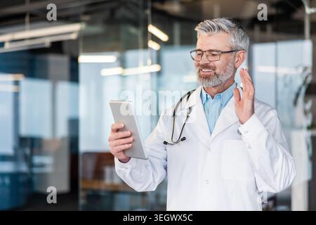 Medico che indossa un camice da laboratorio e uno stetoscopio, sorridente mentre ha una consultazione video online remota su un tablet digitale, offrendo telemedicina per la guarigione Foto Stock