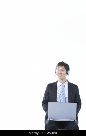 Un giovane uomo si siede con un laptop e le cuffie, sorridendo in un ambiente da studio. Foto Stock