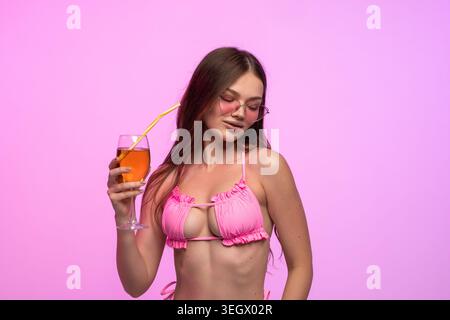 Giovane donna con un top bikini rosa che indossa occhiali da sole a forma di cuore, che regala una bevanda arancione con una paglia per celebrare l'estate Foto Stock